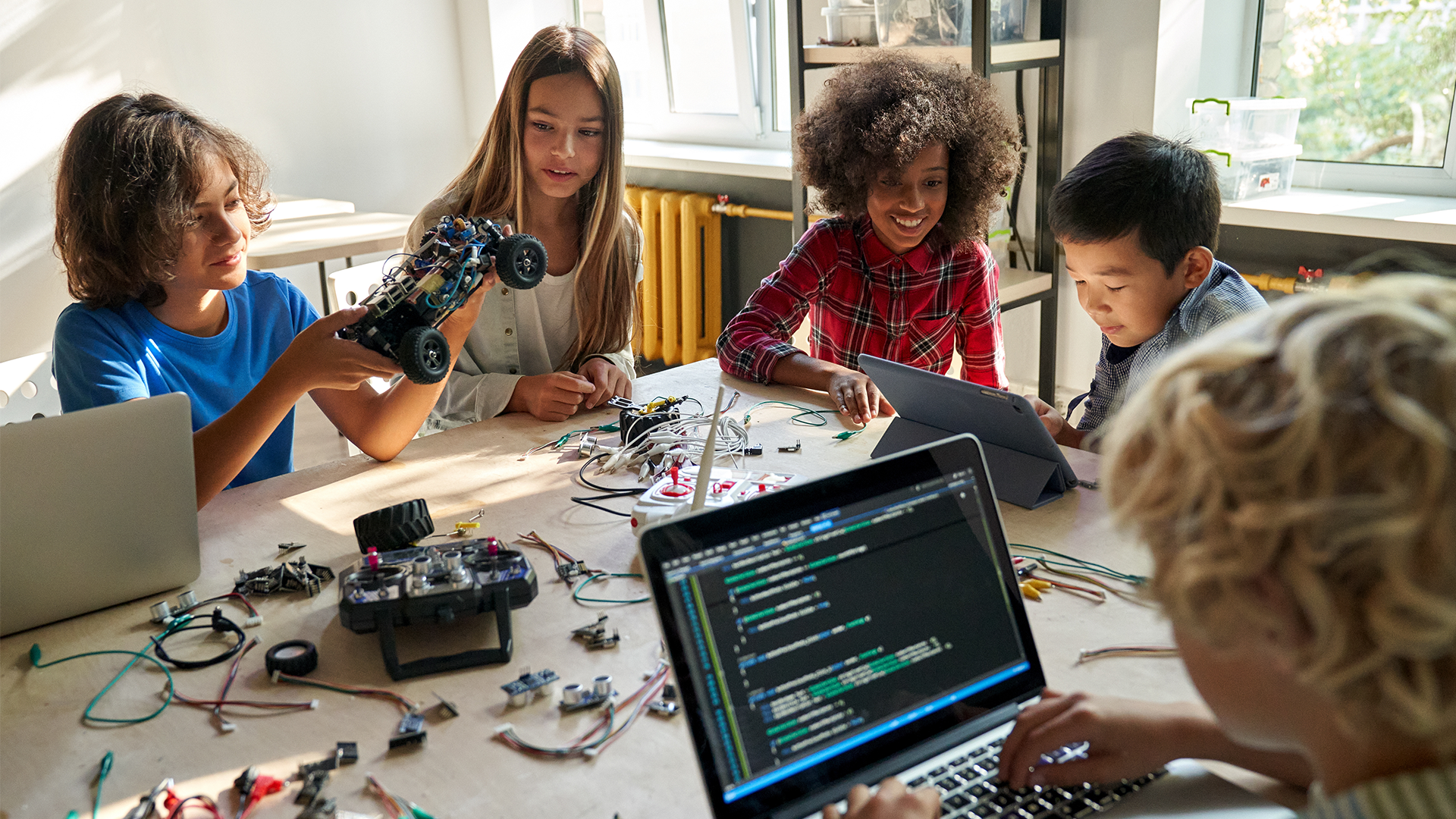 Four children are working together at a table with laptops, circuit boards, wires, and a remote control car. One child holds the car, while others look on and work with electronics in a bright classroom.