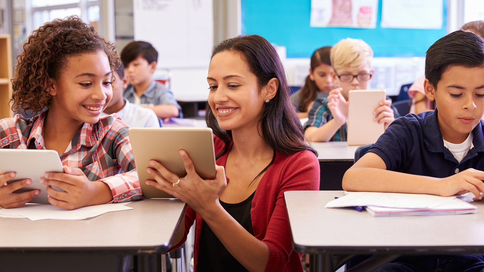A teacher sits at a classroom desk with smiling students, all using tablets. Other students in the background are also engaged with tablets and notebooks. The classroom setting is bright and collaborative.