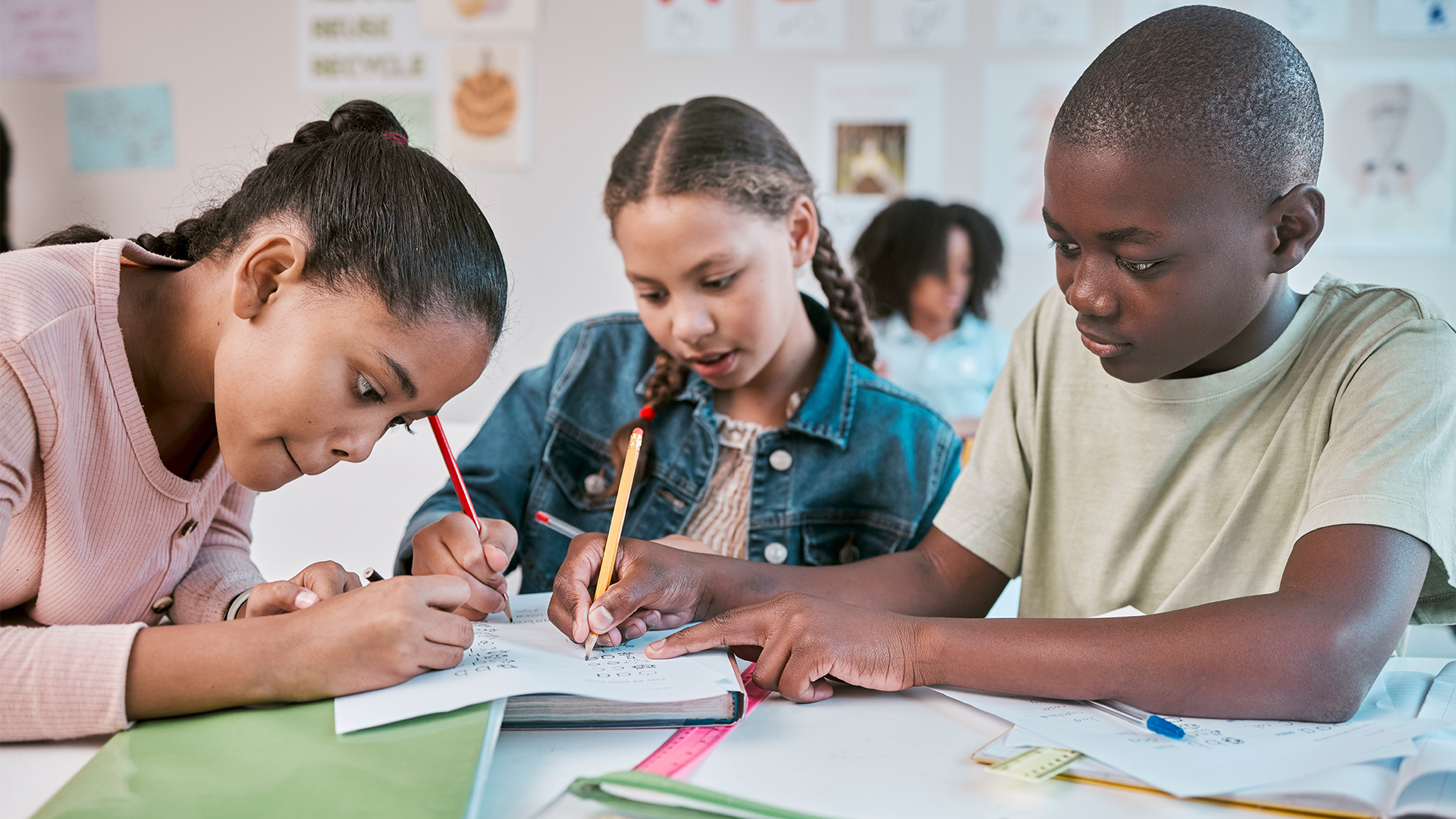 Three children sit at a table in a classroom, working together on school assignments. Two girls and one boy are writing in notebooks, focused and collaborating. Classroom decorations and drawings are visible in the background.