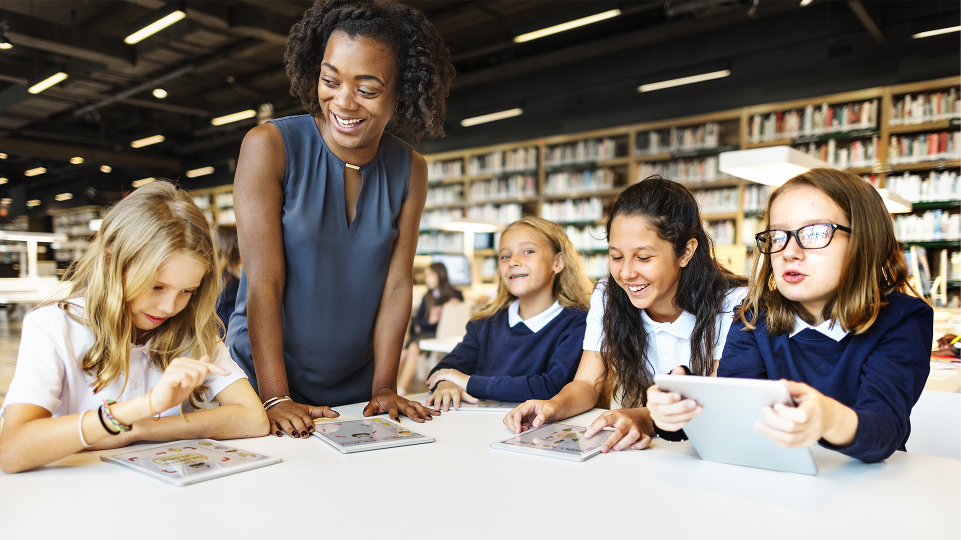 A teacher smiles and engages with a group of five young students using tablets at a table in a library or classroom setting. Shelves filled with books are visible in the background.