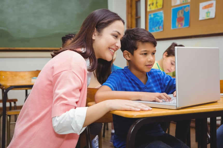 A female teacher looking over the shoulder of a young male (assumed) student looking at a laptop.