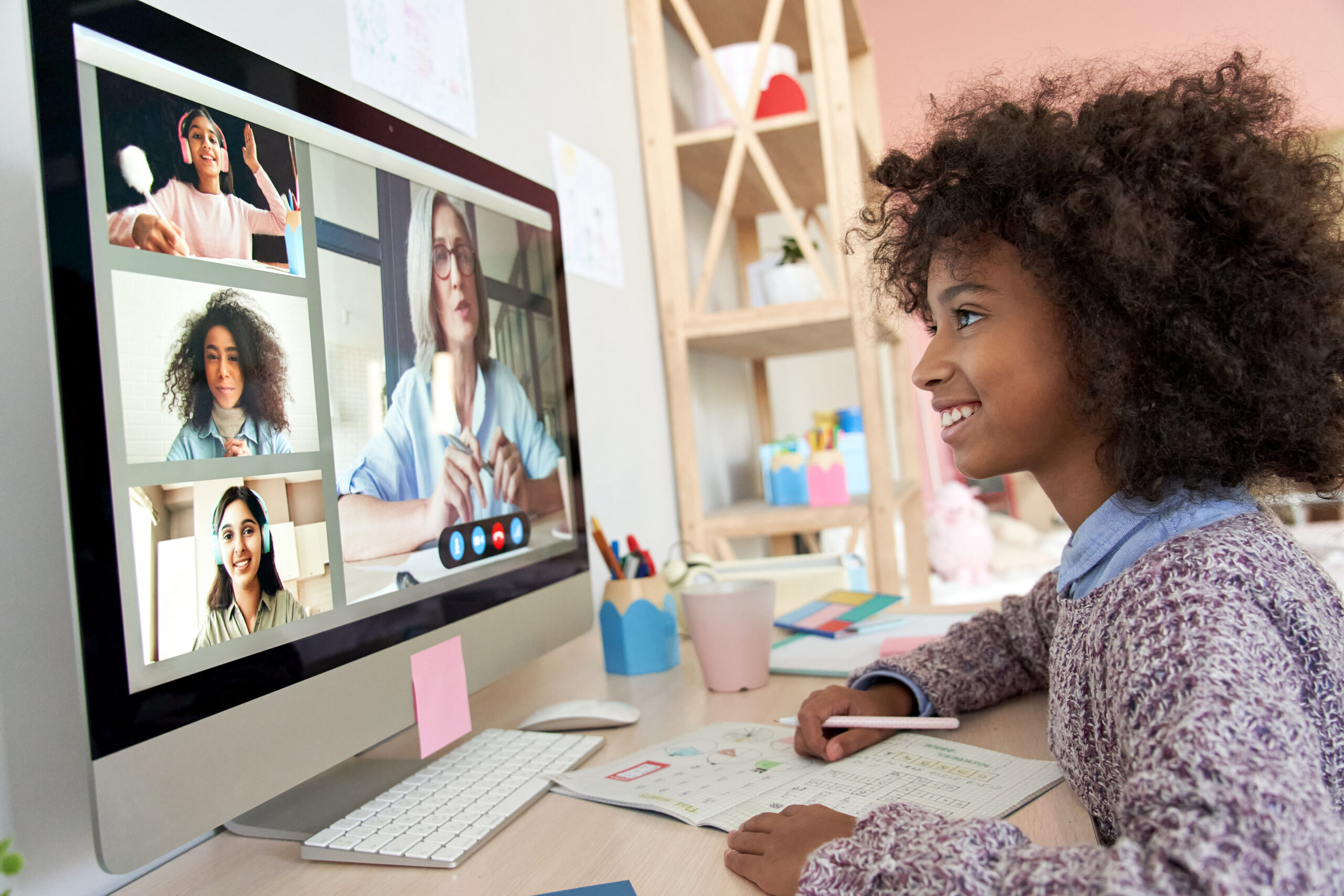 A smiling child sits at a desk with notebooks, attending an online video call on a computer, where four people are visible on the screen. The workspace has colorful supplies and a bookshelf in the background.