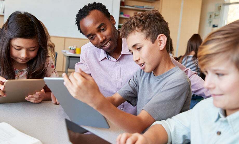 A teacher sits at a table with three students, all looking at tablets, engaged in a classroom activity. The students appear focused and the atmosphere is collaborative and friendly.