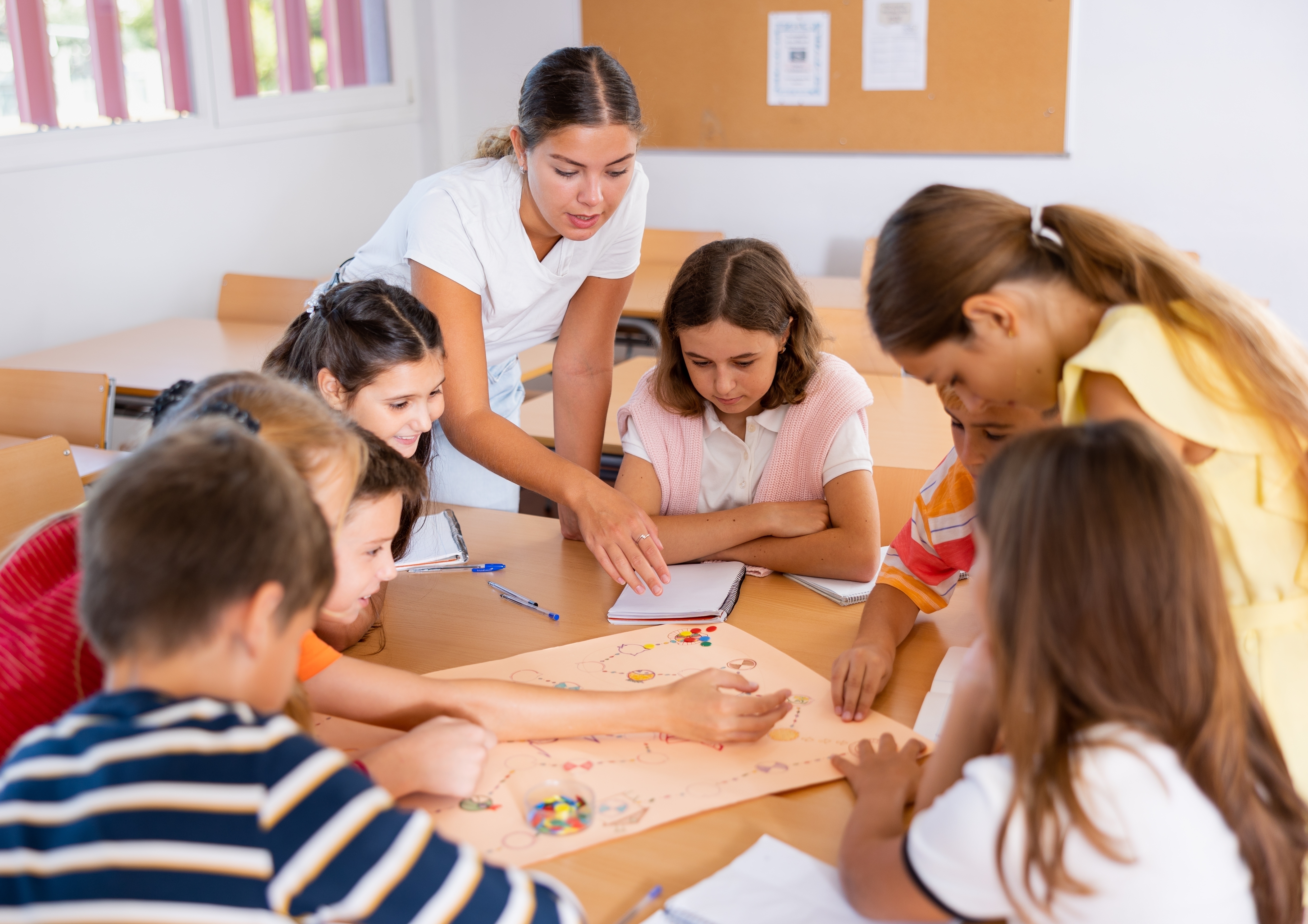 A teacher and a group of children gather around a desk in a classroom, collaborating on a colorful board game or project, with everyone engaged and interacting.
