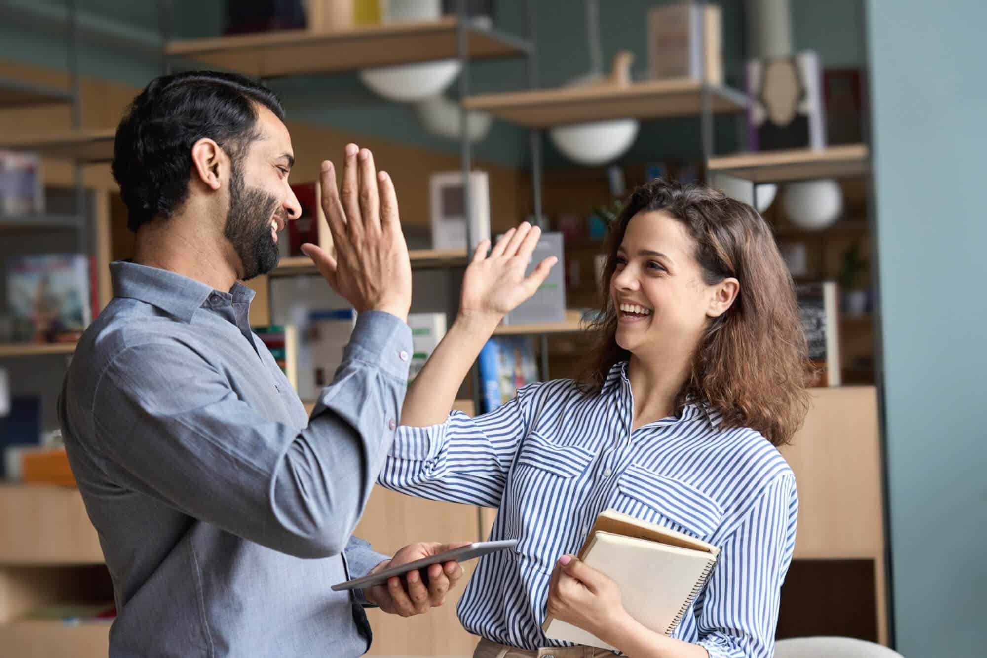 Two coworkers smiling and giving each other a high five in an office setting. One holds a tablet, the other holds a notebook. Shelves with books and decor are in the background.