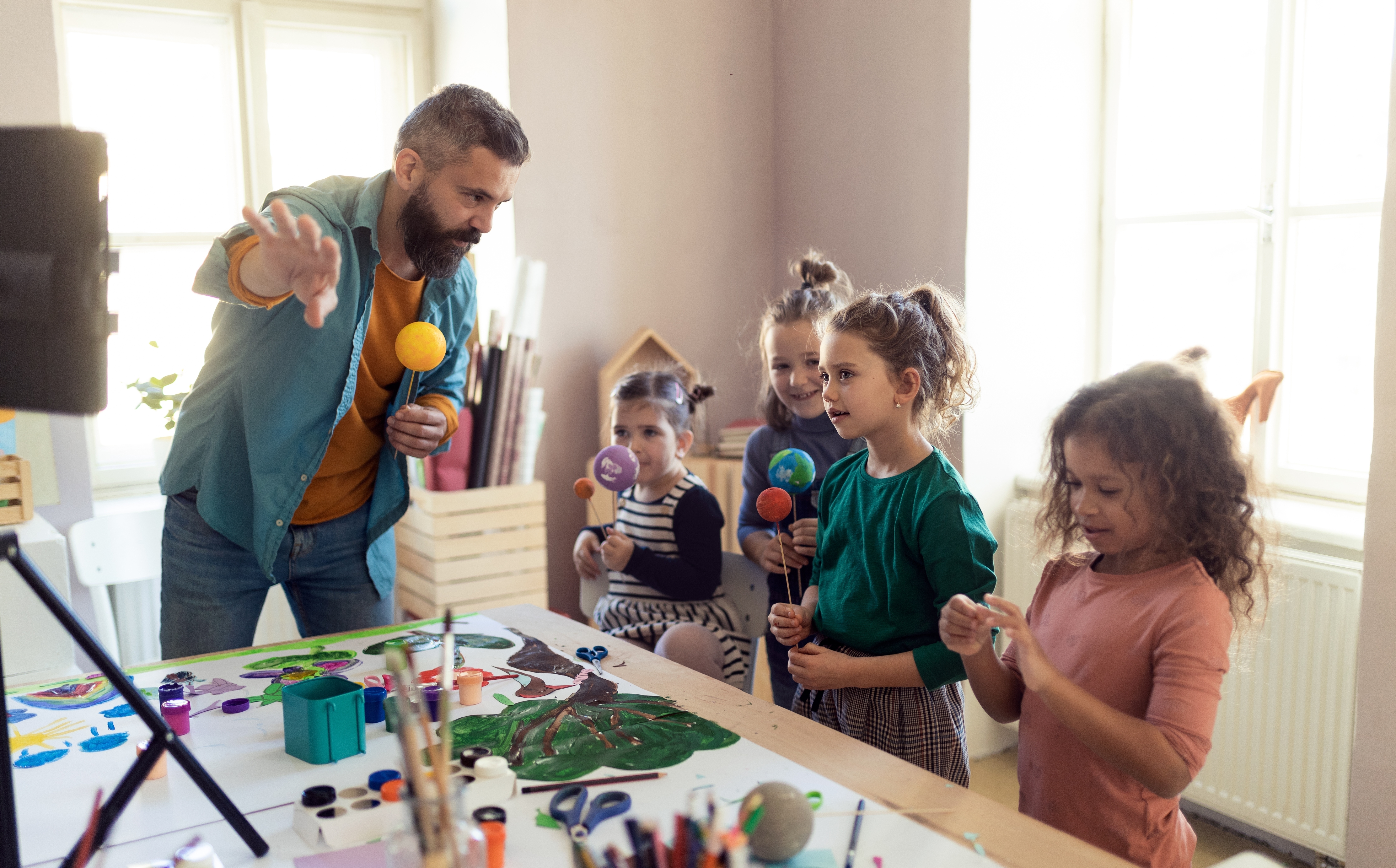 A bearded man is teaching four young girls about the solar system using colorful model planets in a bright classroom with craft supplies on the table.