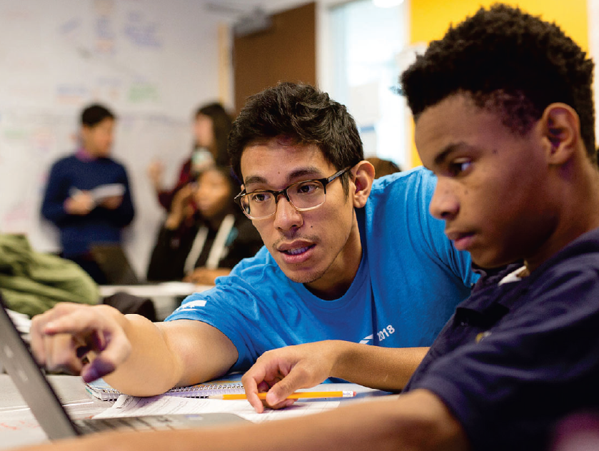 A photograph of two young ethnically-diverse men looking at a laptop screen. One is wearing glasses and a blue shirt and pointing at the screen while the other appears to be a younger student.