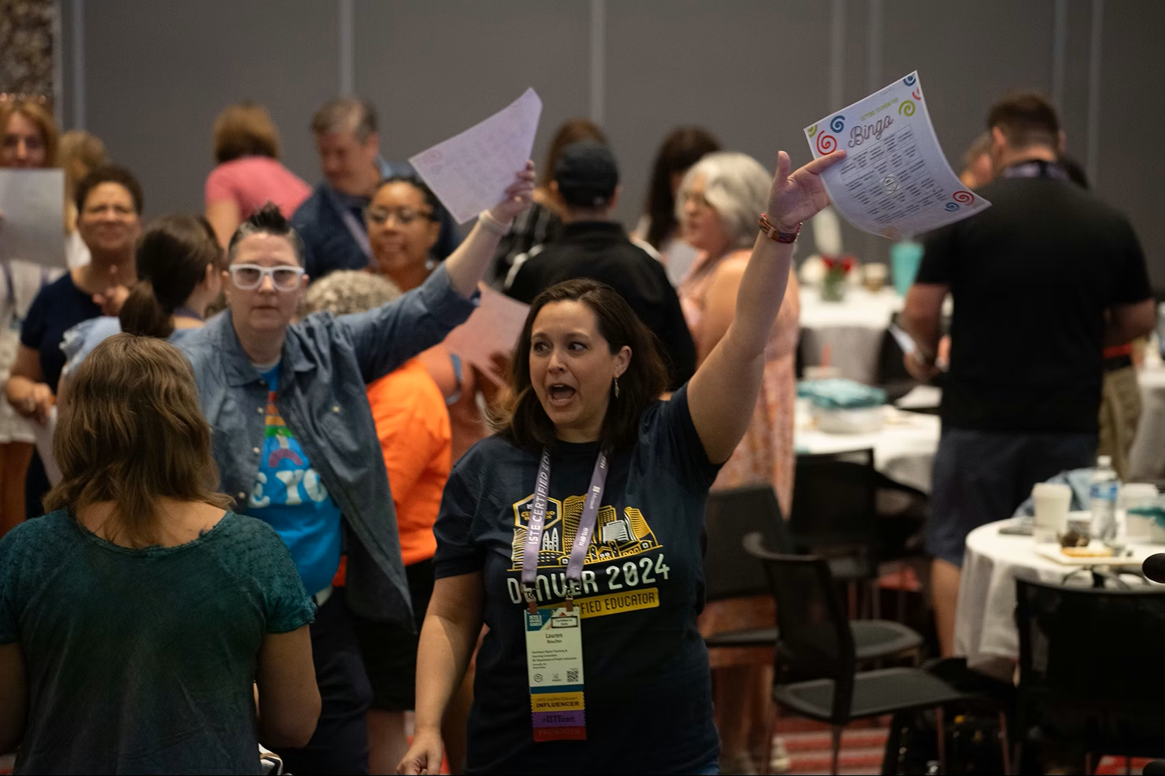 A woman in a conference t-shirt holds up a paper and speaks energetically to a group of people in a busy, well-lit event room with round tables and attendees interacting.