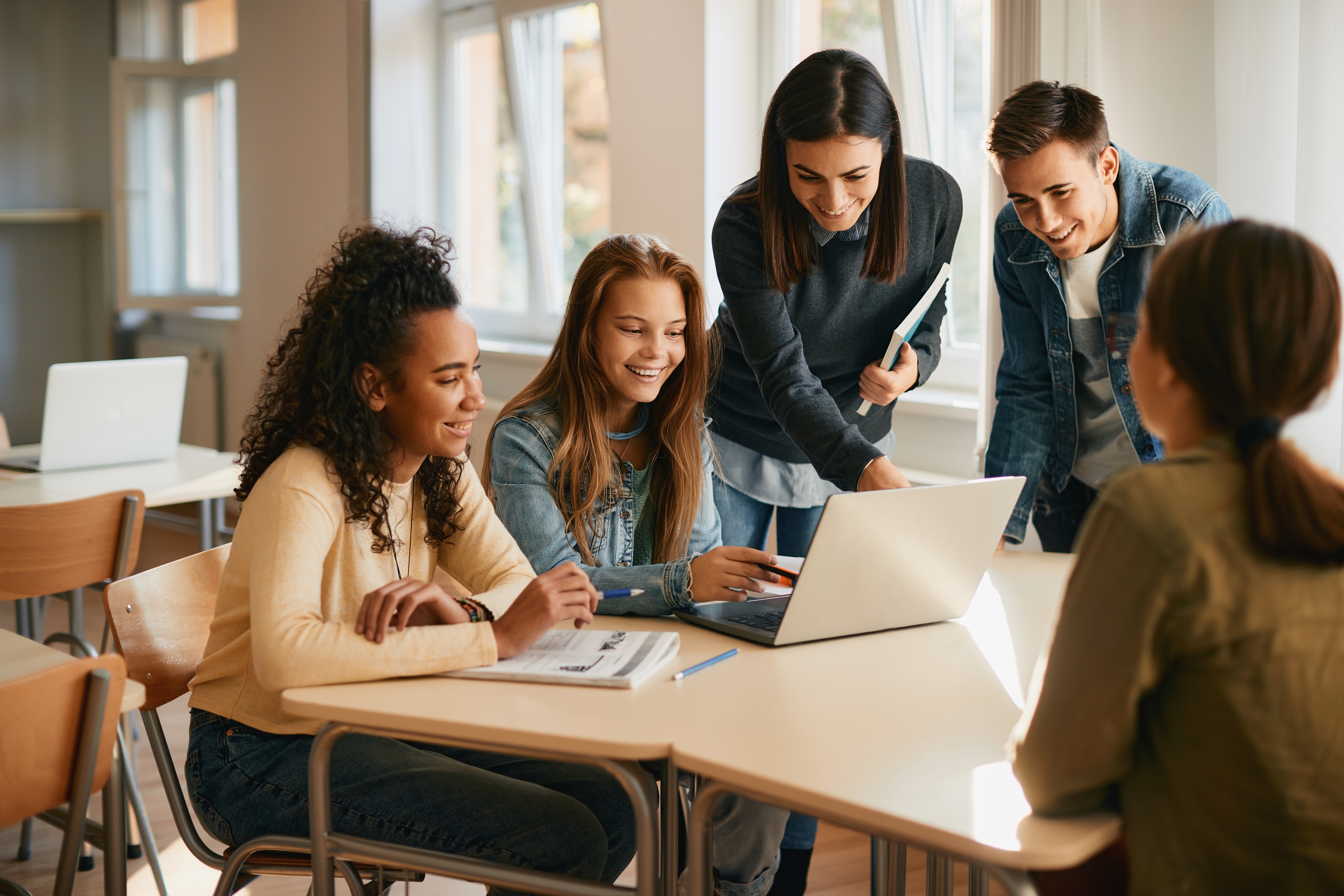 Four young adults sit and stand around a desk in a bright classroom, smiling and looking at a laptop screen together. Open books and notebooks are on the desk, suggesting a collaborative study session.
