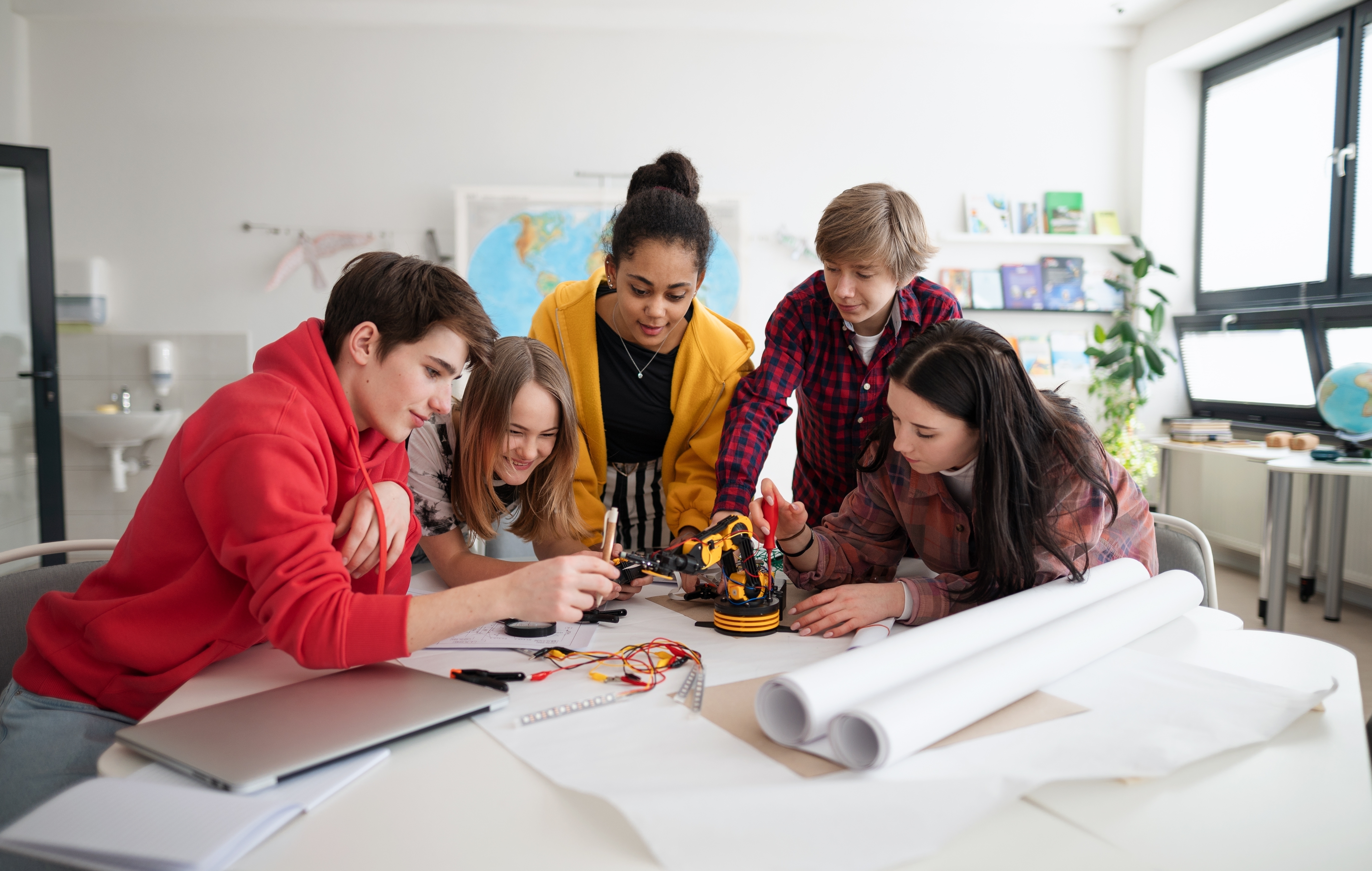 A group of five teenagers gathered around a table, collaborating on building a robotic arm. Blueprints, wires, and a laptop are on the table, suggesting a STEM or robotics project in a bright classroom.
